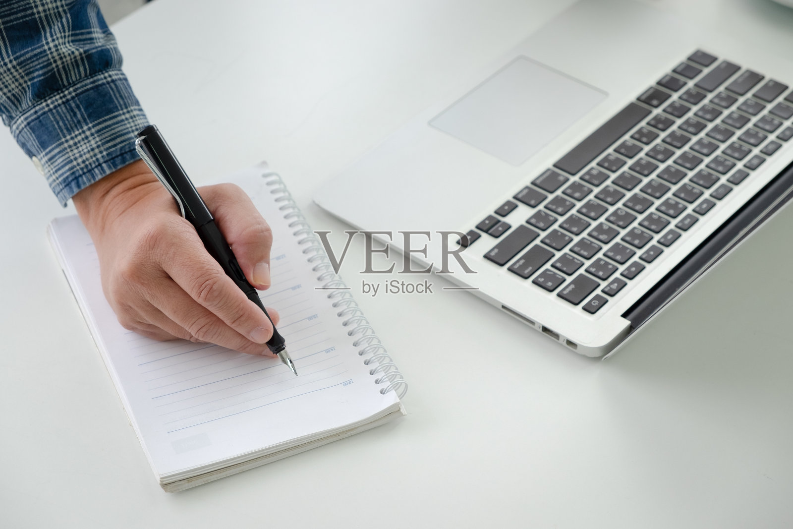商人的手拿着一支黑色的笔，在一张白色的桌子上的笔记本电脑附近写便条。照片摄影图片