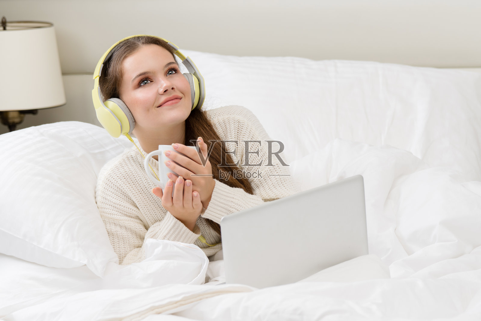 美丽迷人的年轻白人女人黑发早上在白色的床上与杯子在手提电脑看脸微笑梦想看在耳机听音乐照片摄影图片