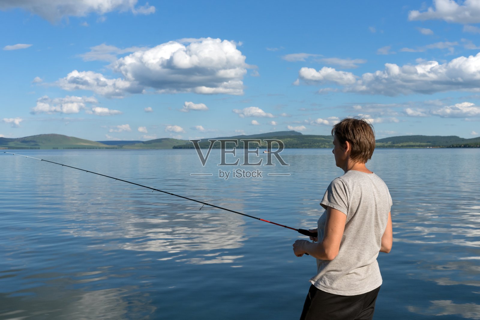 在多山的海岸边，一个女渔夫在一个蓝色的湖里钓鱼。照片摄影图片