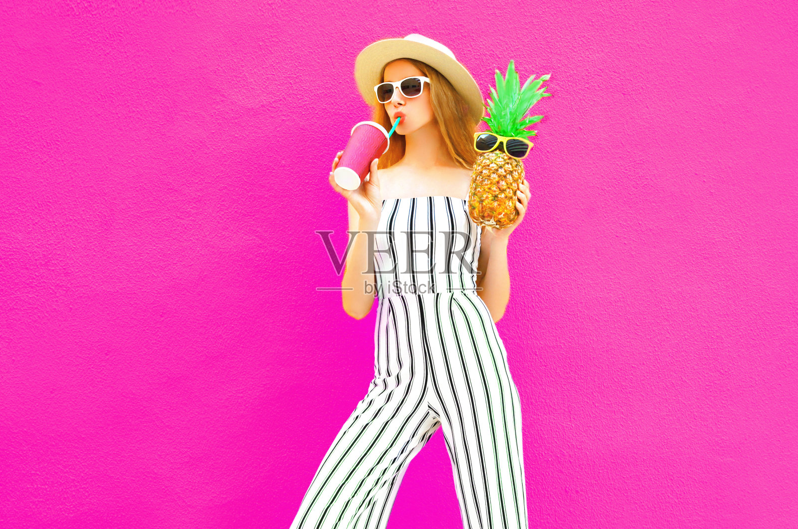 肖像时尚的女人喝果汁拿菠萝在夏季圆帽，白色条纹连衣裤上彩色的粉红色墙壁背景照片摄影图片