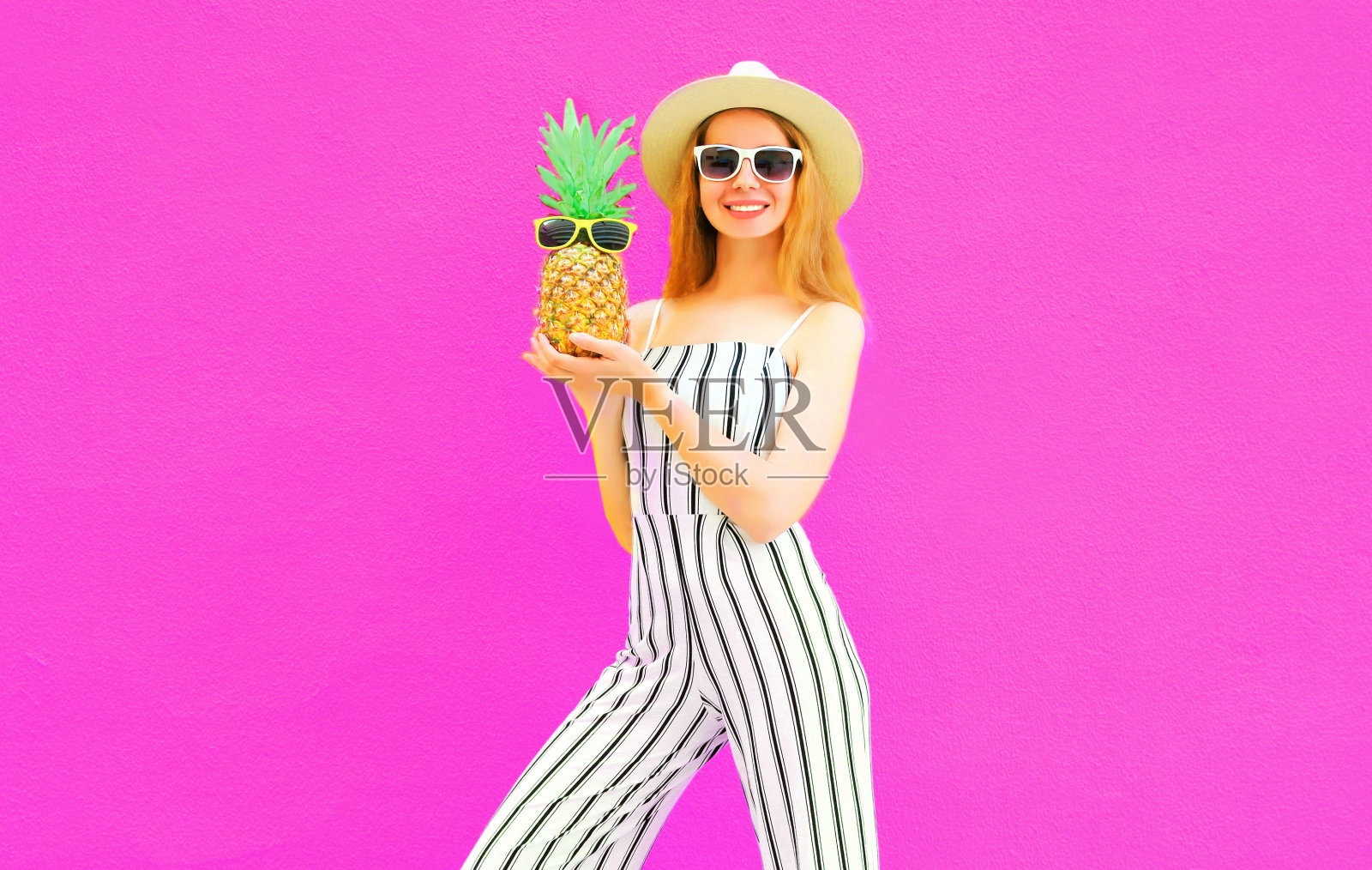 时尚快乐微笑的女人抱着菠萝穿着条纹连衣裤，夏天圆草帽上五颜六色的粉红色背景照片摄影图片