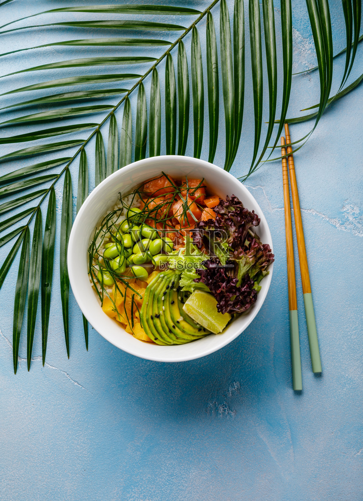 三文鱼戳碗生鱼沙拉是亚洲流行的食物，以大豆、毛豆、大米、鳄梨、热带水果和生菜在碗里，热带的叶子和蓝色的背景照片摄影图片