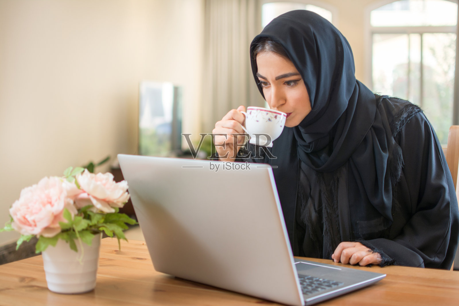 年轻的阿拉伯妇女喝着咖啡，在家里用笔记本电脑看新闻照片摄影图片