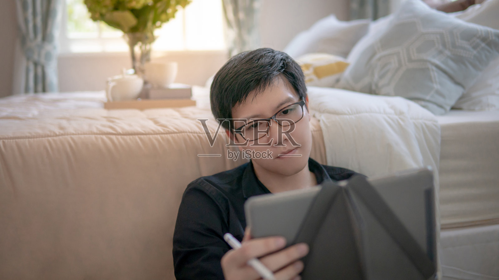 在舒适的卧室里，年轻的亚洲商人坐在床边使用数字平板电脑和笔。具有现代电子小玩意概念的家居生活方式照片摄影图片