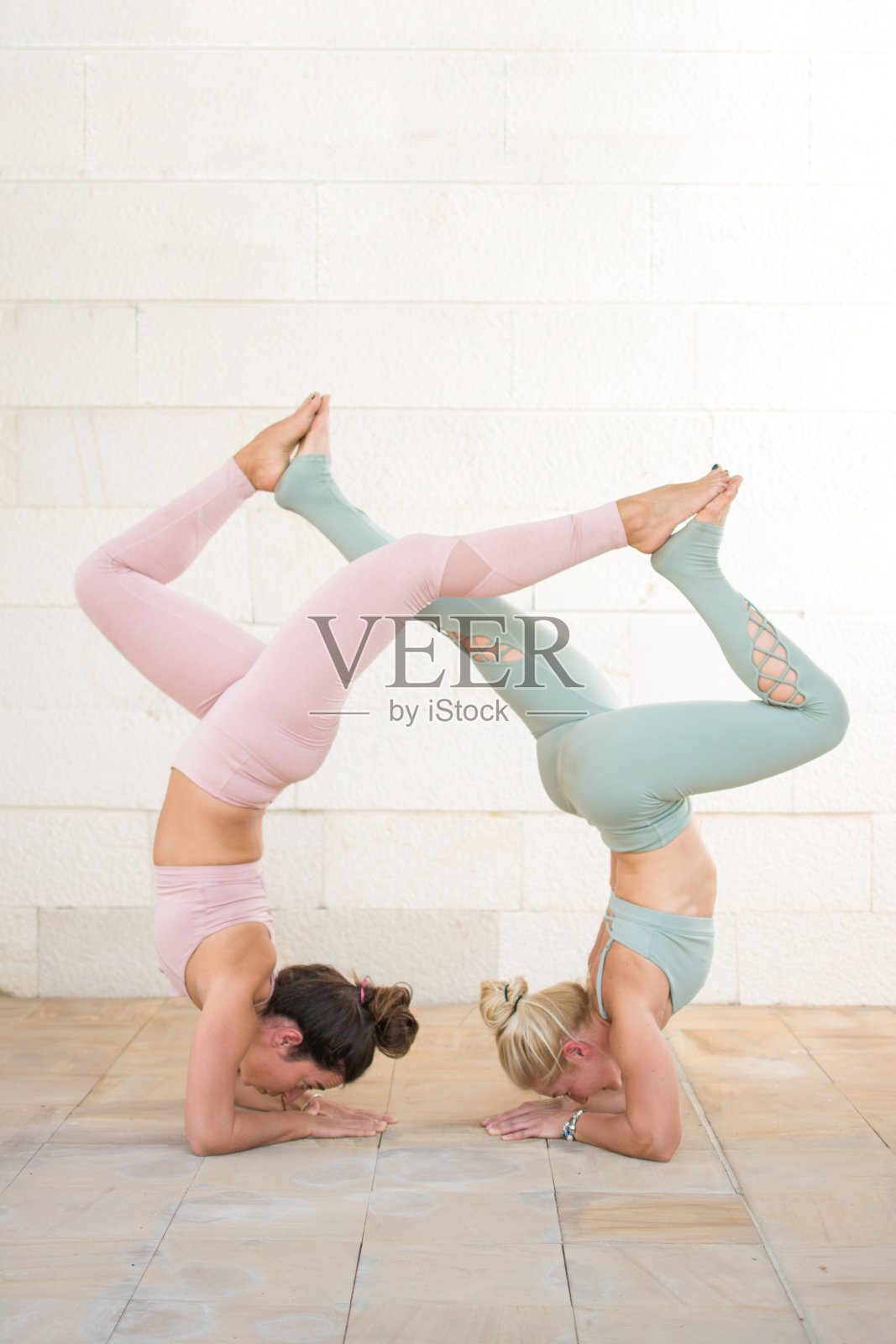 两个运动女性一起进行倒立瑜伽练习。健身女孩做倒立练习，双腿互相触碰在白色的墙壁上。照片摄影图片