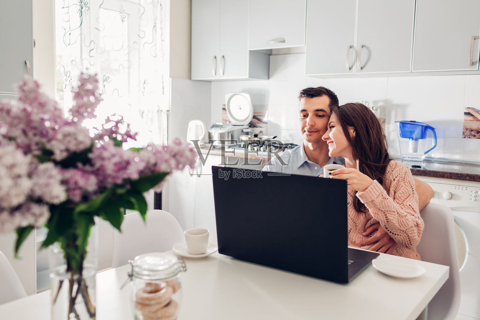 幸福的年轻夫妇在现代厨房里用笔记本电脑吃早餐。年轻男女喝着咖啡，拥抱着照片摄影图片