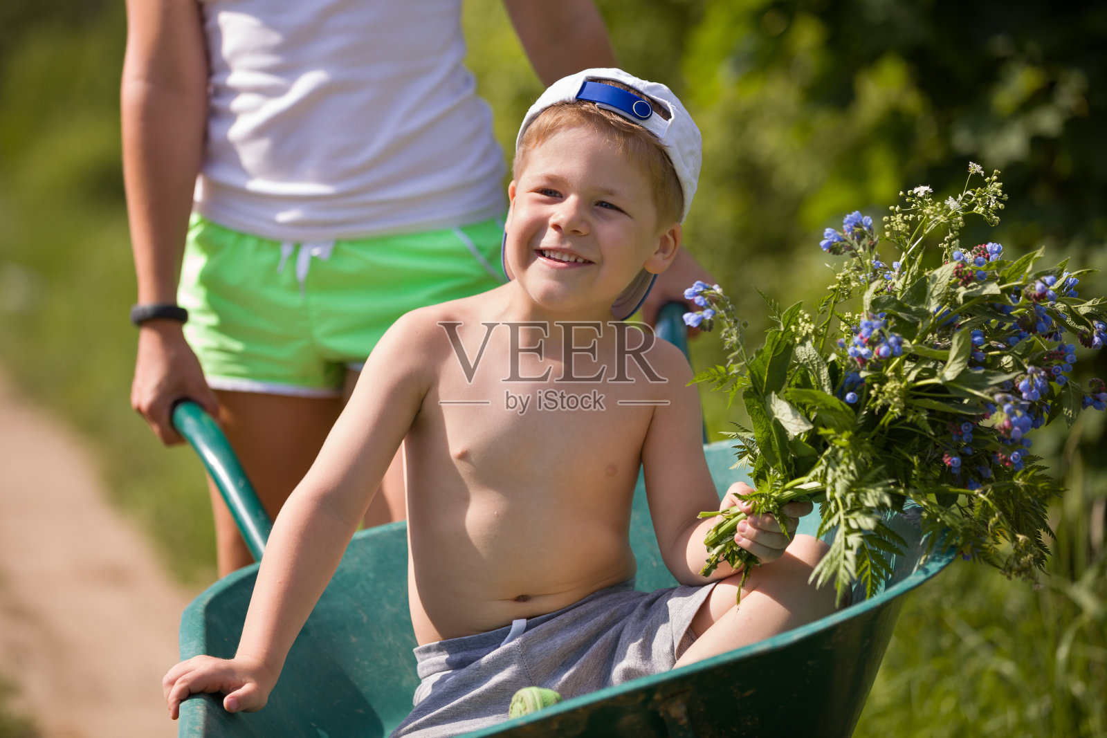可爱的快乐男孩的肖像抱着一束野花，坐在一个花园手推车。在一个阳光明媚的日子里，年轻的母亲拉着一个微笑的孩子。的生活方式。照片摄影图片