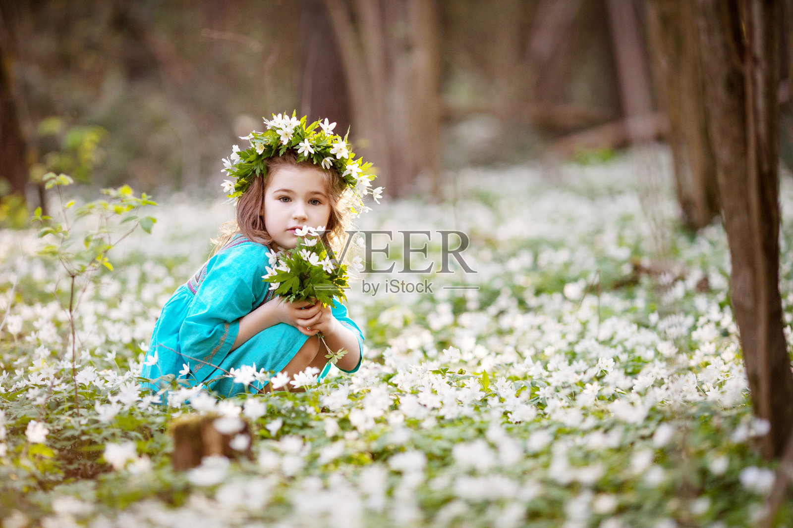 美丽的小女孩穿着蓝色的裙子走在春天的树林里。一个头上戴着花环的漂亮女孩的肖像。夏天，孩子们在户外玩耍。复活节的时间。可爱的园丁种植雪滴。照片摄影图片