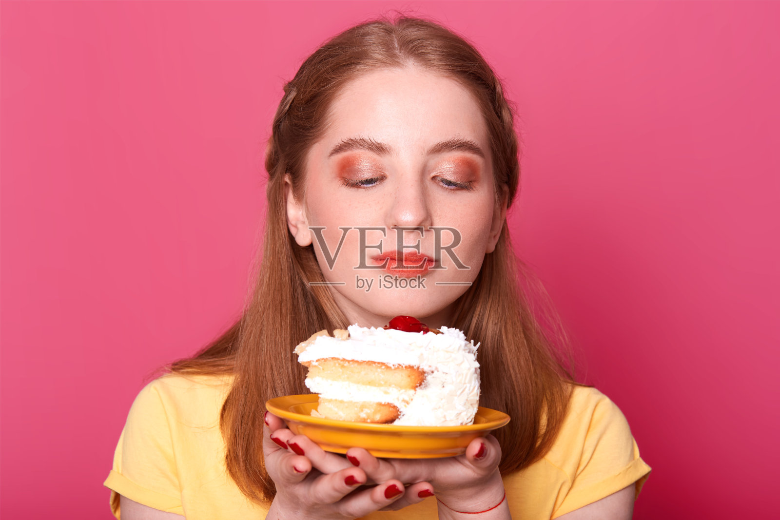 漂亮的年轻女孩的肖像，看着盘子与一块生日蛋糕孤立在粉红色的背景，想要吃美味的甜点，穿着黄色的t恤，有完美的发型，在工作室摆姿势。照片摄影图片