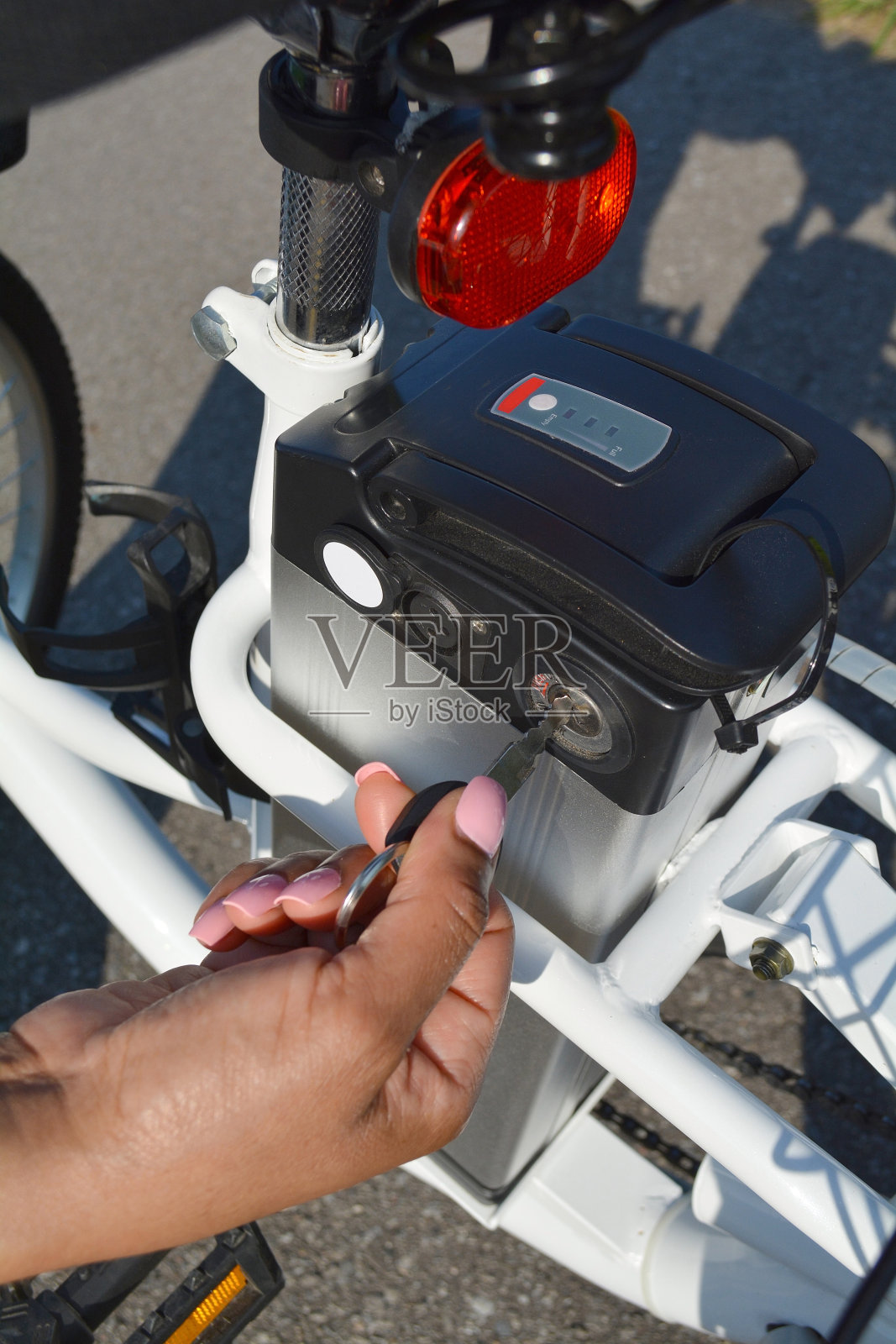 女子自行车手握着电动自行车电池包的钥匙。女开关电动自行车与钥匙在公园在阳光明媚的夏天。近距离射杀。照片摄影图片