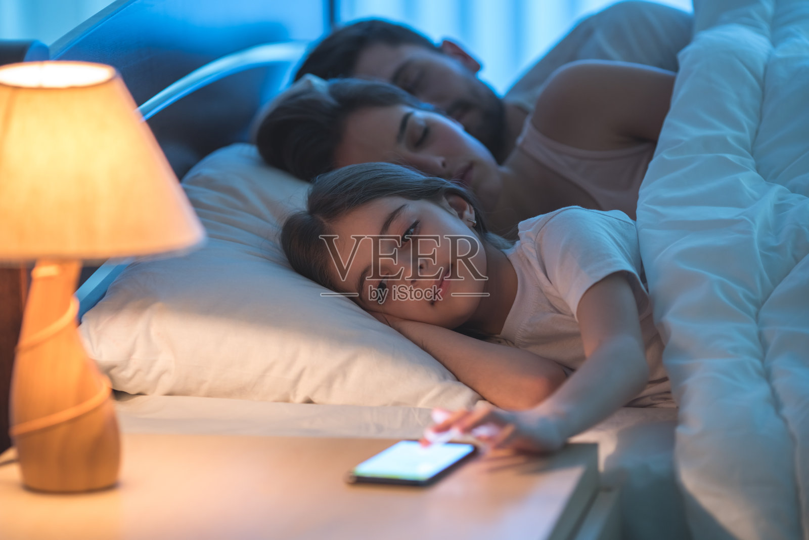 女孩在熟睡的父母旁边打电话。晚上的时间照片摄影图片