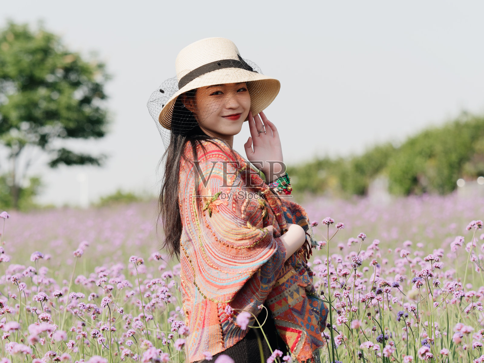 身穿黑色马鞭草背心、披着彩色披肩、戴着草帽的美丽女子，在紫色的马鞭草花海中摆姿势，一头乌黑长发的迷人中国女孩在户外享受闲暇时光。照片摄影图片