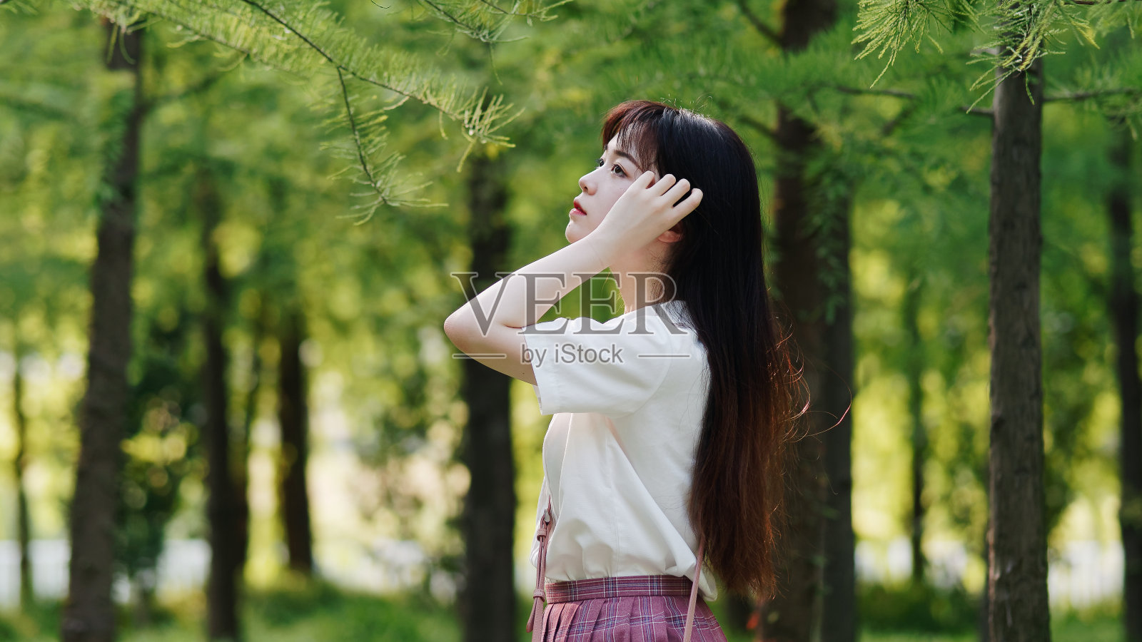 美丽的中国女孩的肖像在白色的衬衫和粉红色的裙子看与手的头发在阳光灿烂的夏天森林，呼吸新鲜的空气和享受自然。美丽，生活方式和夏季概念。照片摄影图片