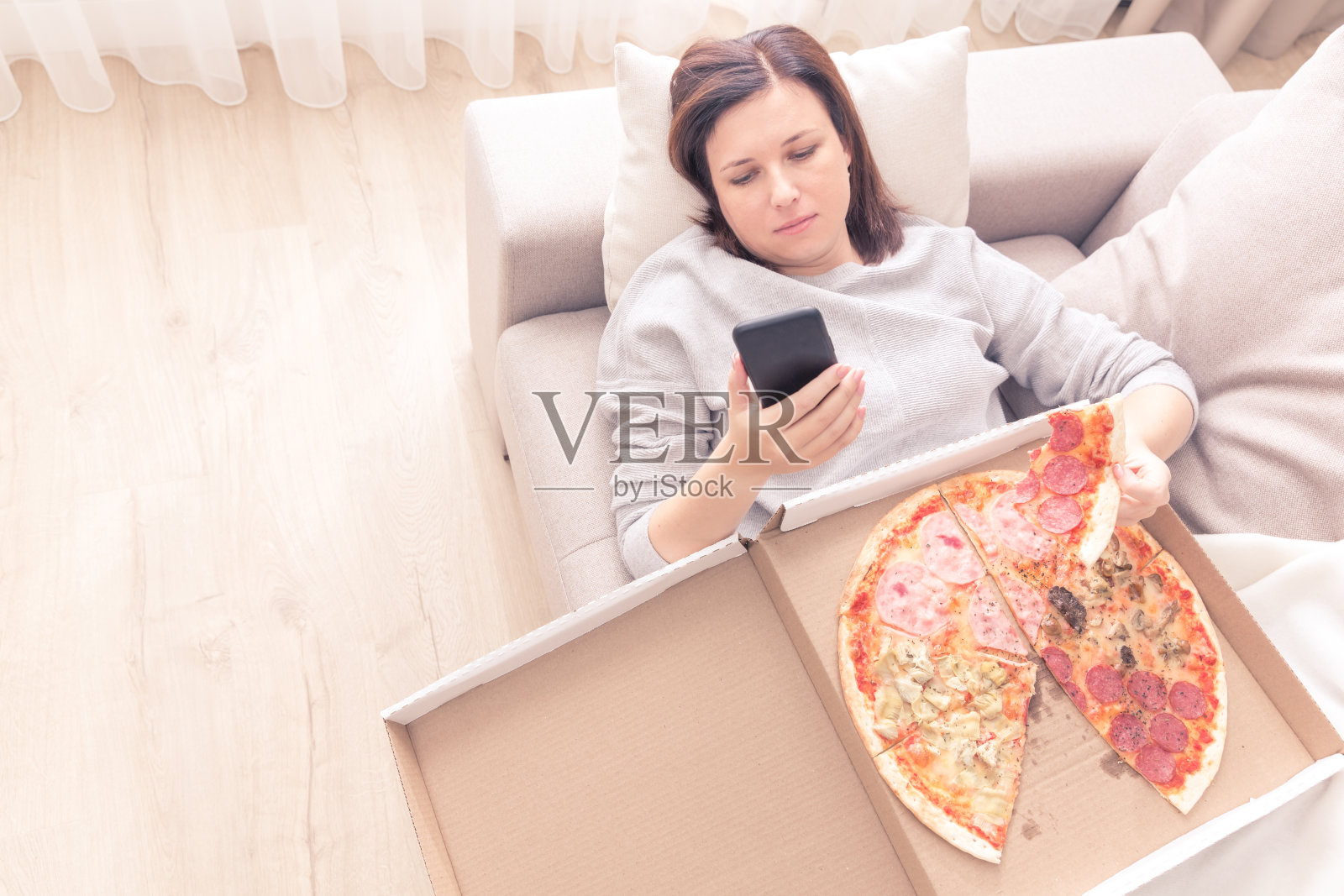 郁闷的女人躺在家里的沙发上吃着披萨拿着电话，暖暖的黄色调照片摄影图片