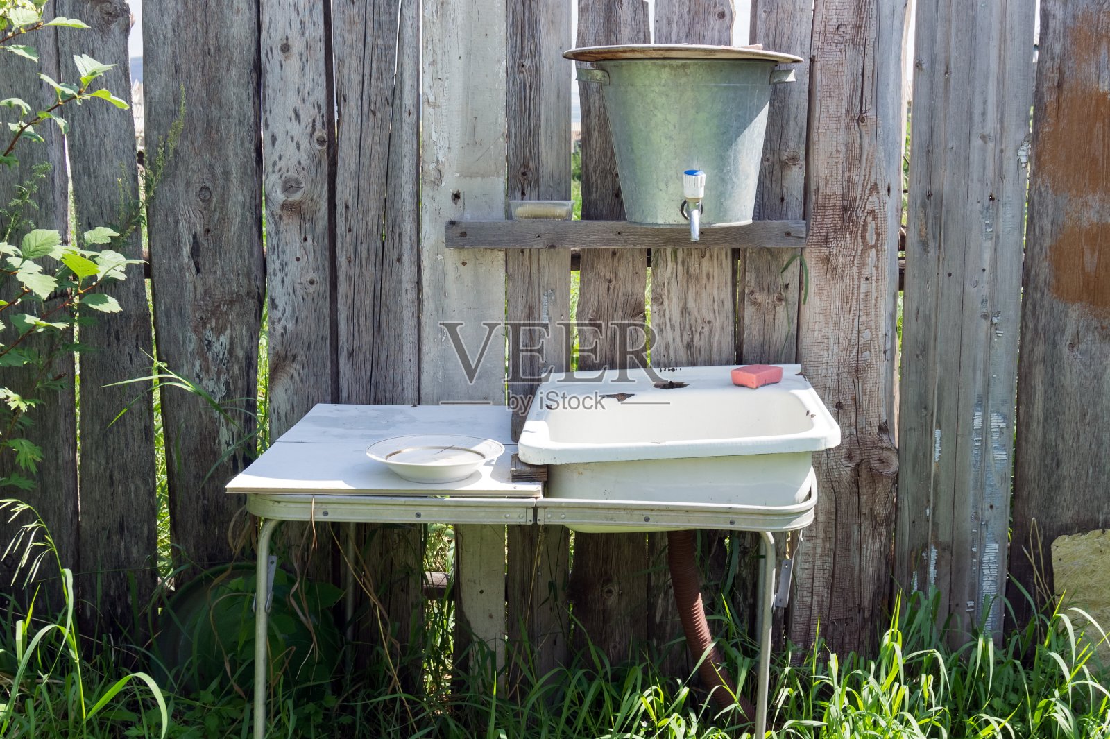 夏天，木栅栏旁的绿草地上放着一个带水槽的质朴金属盥洗架。照片摄影图片
