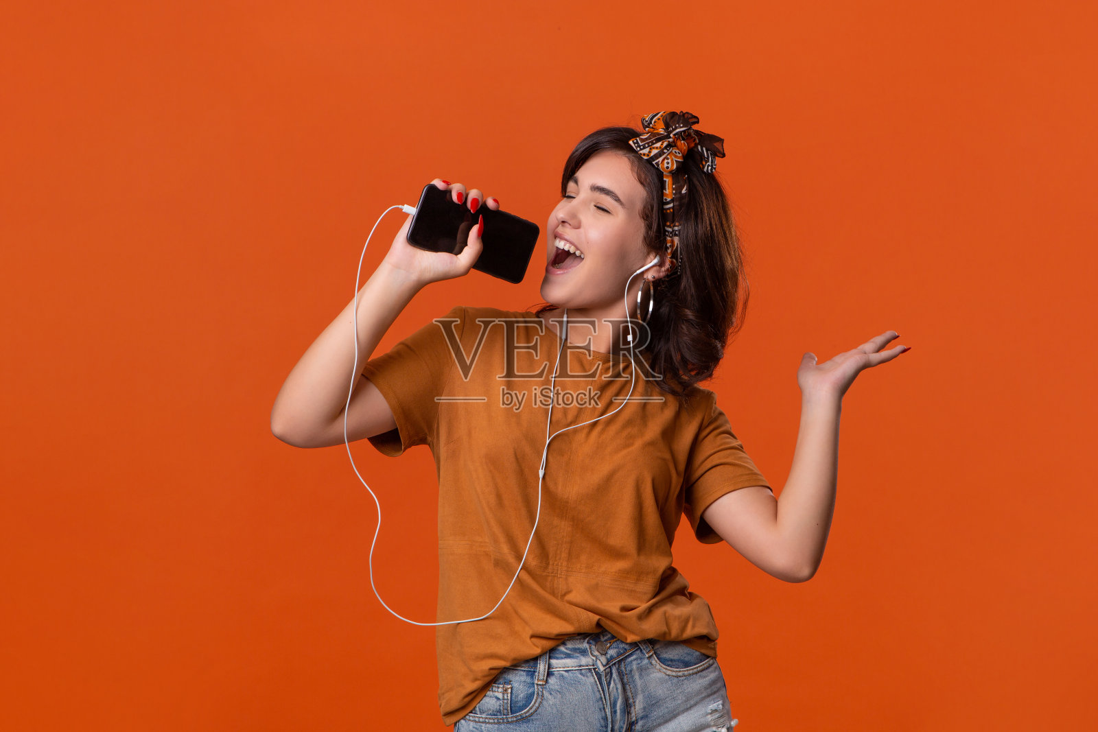 漂亮的黑发女子在一件t恤和美丽的头带跳舞听音乐与有线耳机隔离在橙色的背景。享受生活照片摄影图片