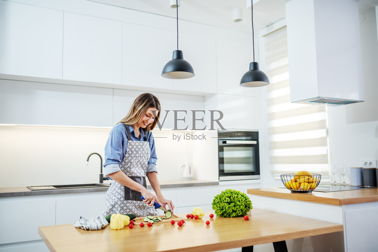 年轻幸福的白人女人穿着围裙站在厨房切黄瓜。厨房台上放着各种蔬菜。健康饮食的概念。照片摄影图片