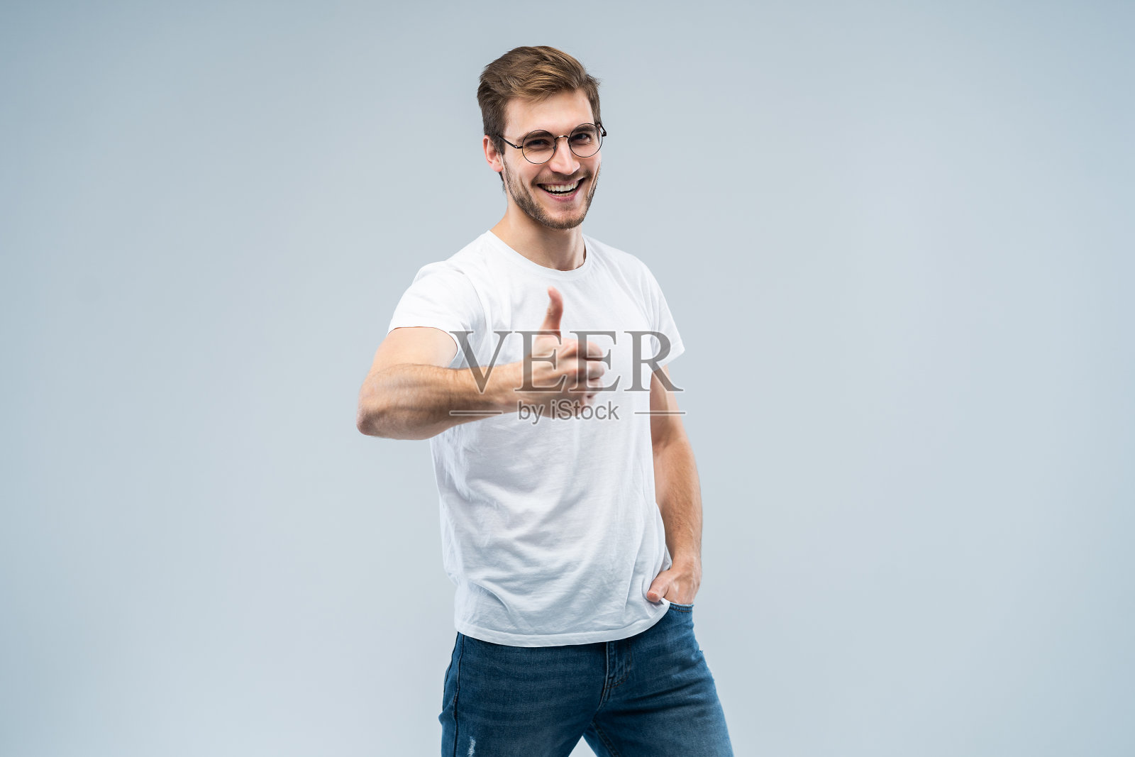 一个有魅力的男人在相机上竖起大拇指，在灰色的背景下露出幸福的微笑。照片摄影图片
