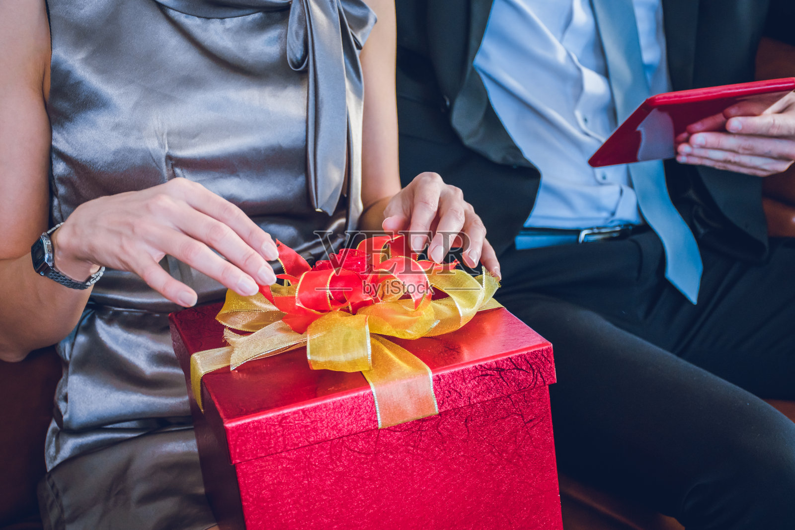 一个女人坐在棕色的沙发上，房间里有一面白色的墙。她手里拿着一个红色的礼品盒，上面写着新年快乐，情人节。一个拿着红色智能手机的男人。照片摄影图片