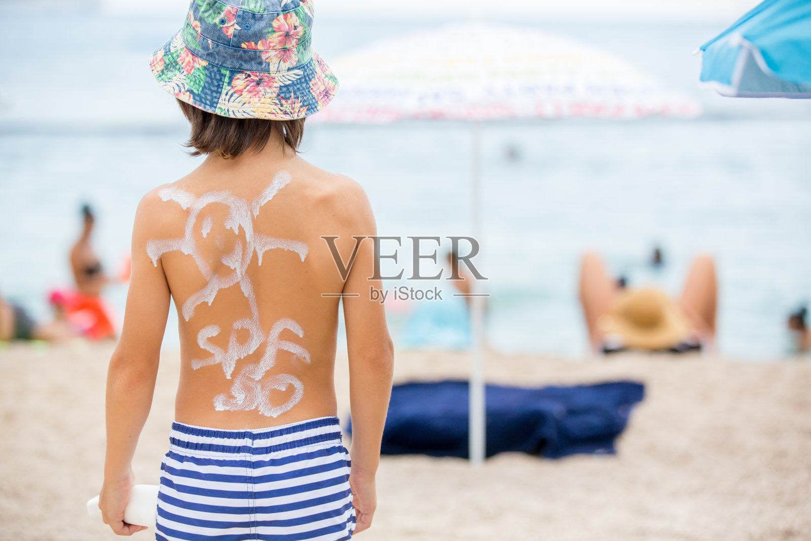 可爱的学龄前男孩，拿着一个背上涂了防晒霜的防晒霜瓶子，准备迎接刺眼的太阳照片摄影图片