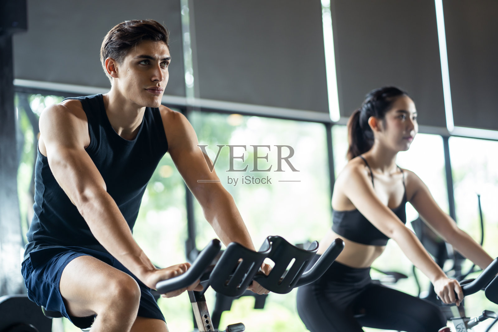 英俊的白人运动员和亚洲可爱的女孩在现代健身房或健身俱乐部。他们骑着自行车。健身房和健身的良好健康理念。照片摄影图片