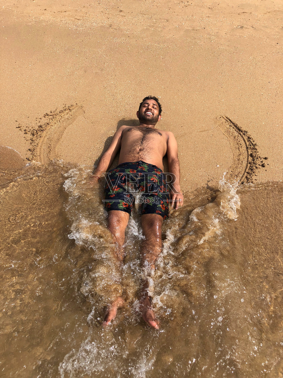 赤裸上身的印度年轻男子在沙滩上做雪天使，在海边度假时穿着泳裤晒太阳，涂上防晒霜，胸部和腿部多毛，手臂和腿部活动照片摄影图片