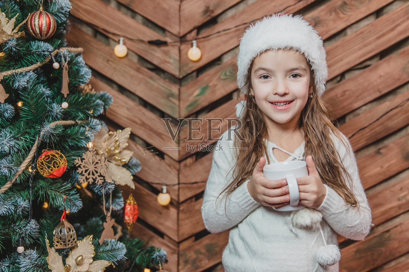 可爱微笑的女孩坐在舒适的家里的特写照片，拿着一杯热巧克力在圣诞树上的木制背景。照片摄影图片