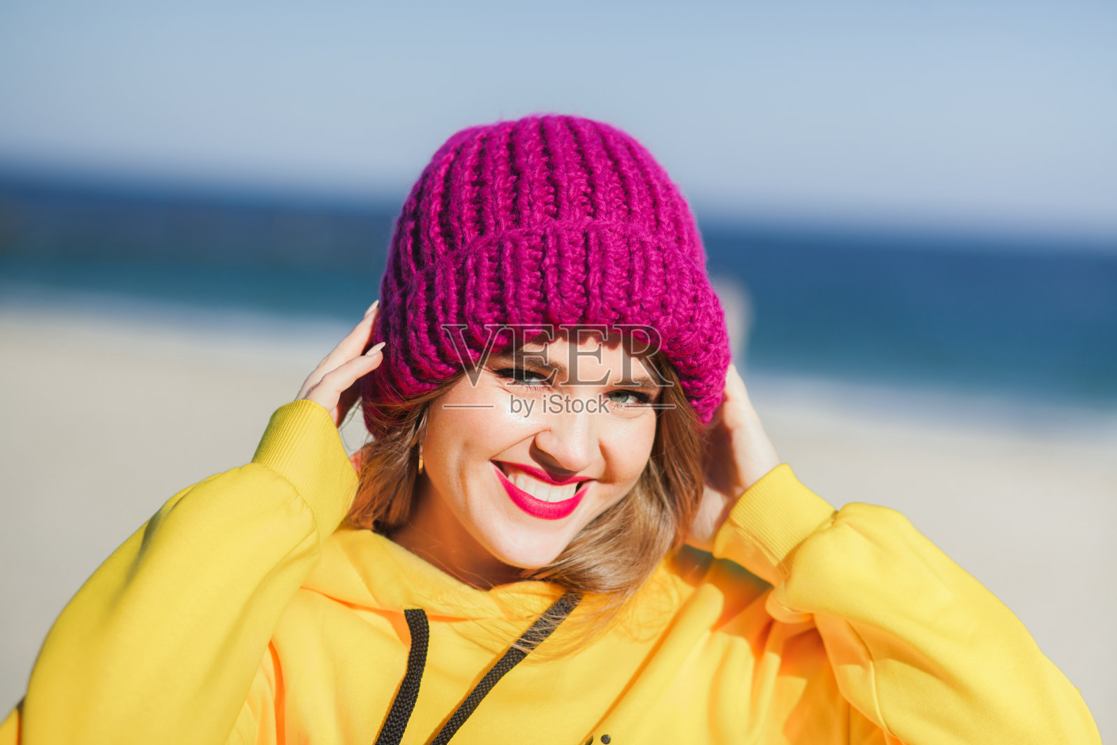 年轻漂亮的女人在黄色的运动衫和明亮的紫色针织帽在沙滩上在阳光明媚的日子照片摄影图片