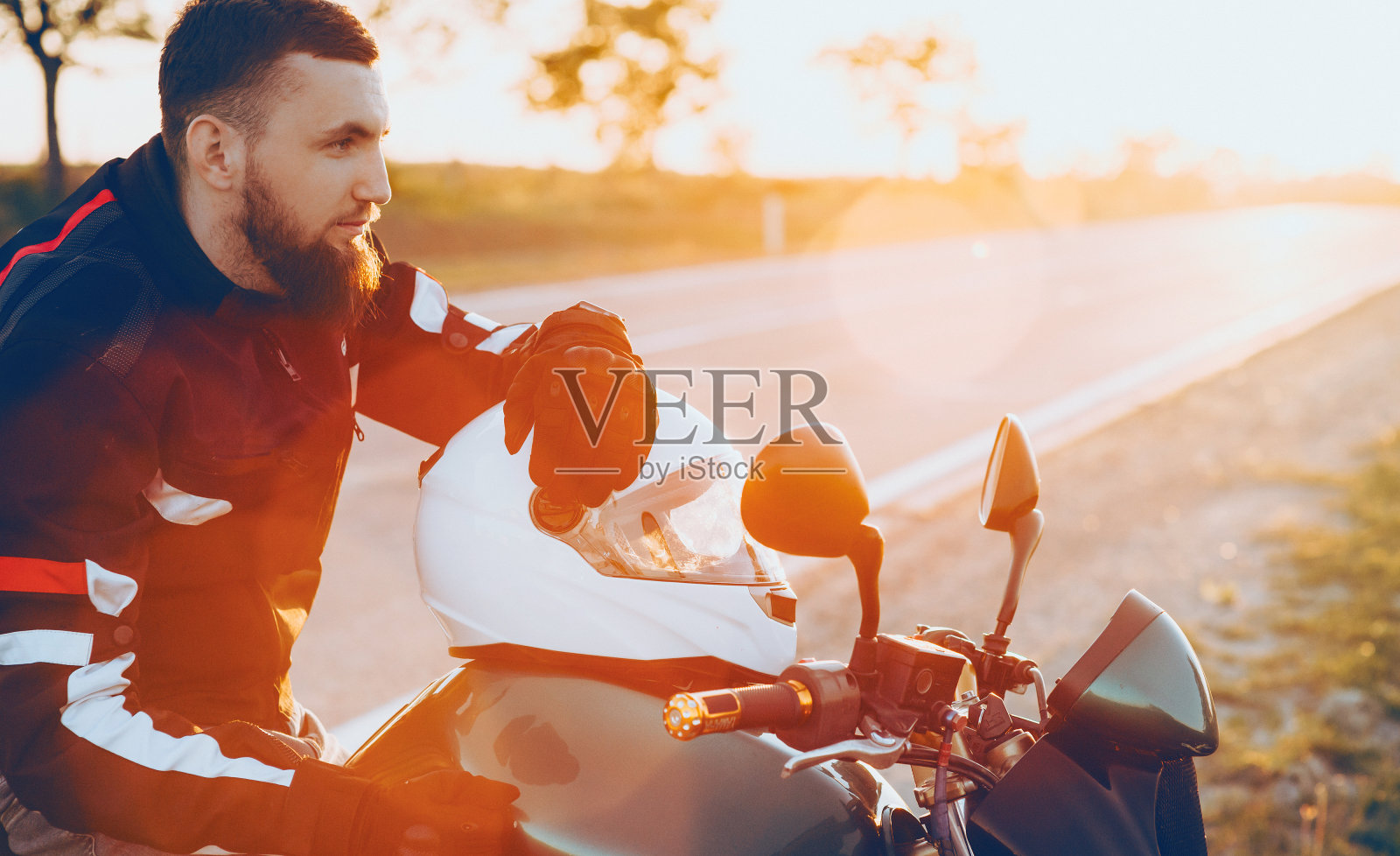 自信的白种人大胡子坐在他的摩托车上，把他的手放在白色的头盔上，而太阳正在温暖他照片摄影图片