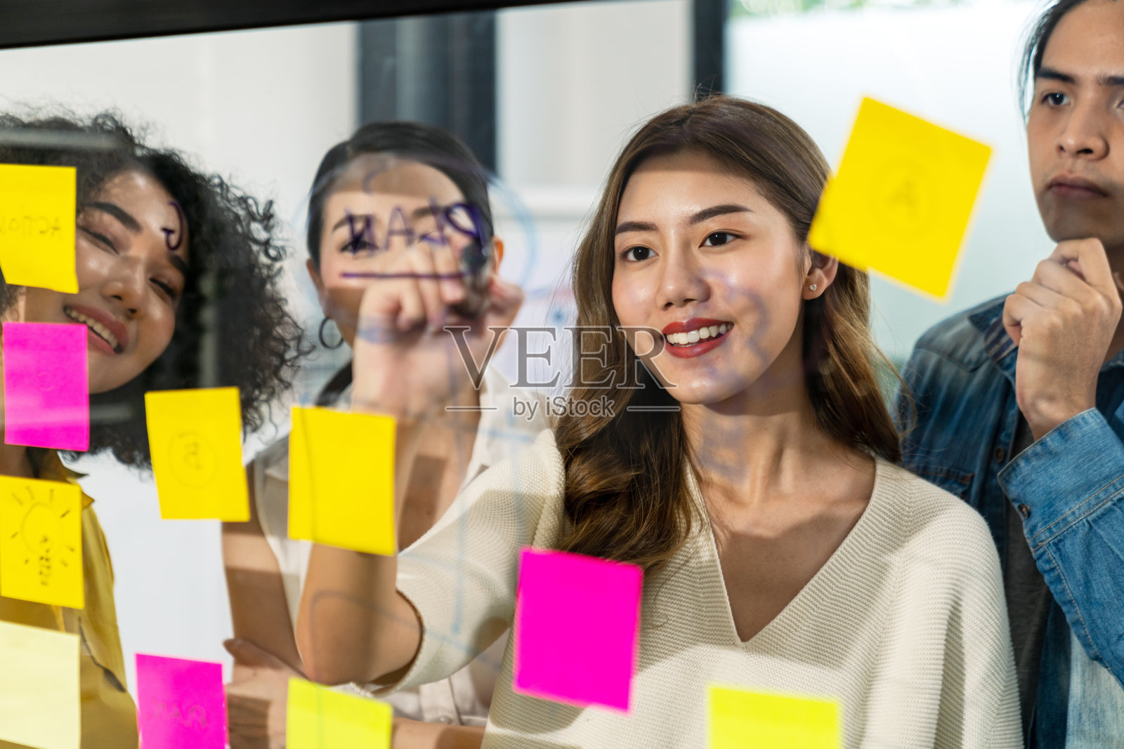 亚洲富有创意的女性微笑着在办公室开会，用便签来分享想法和计划项目。一组员工在玻璃墙和粘纸上集思广益。头脑风暴的概念。照片摄影图片