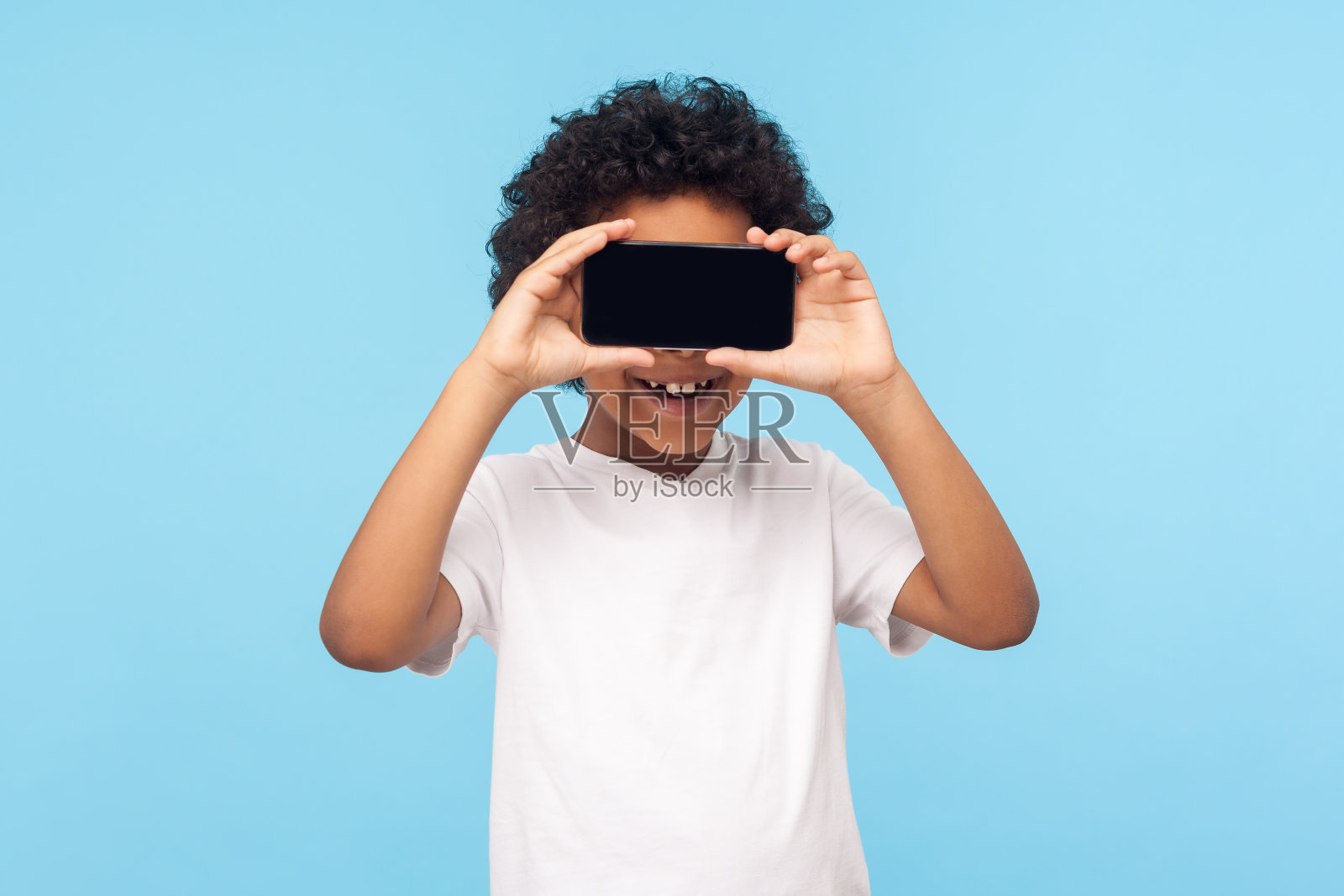 积极可爱快乐的小男孩卷着头发，用手机遮住眼睛，微笑着照片摄影图片