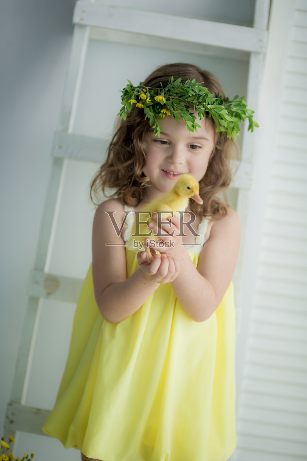 一个头上戴着花环，穿着黄色衣服的可爱美丽的女孩，抱着一只黄色的小鸭子，背靠一堵白色的墙照片摄影图片