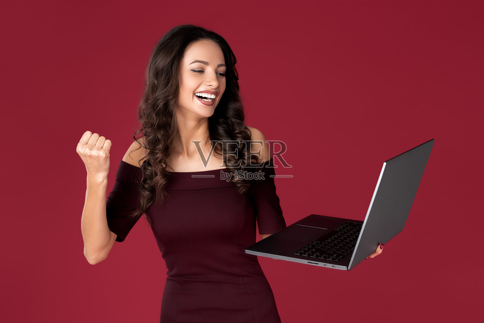 漂亮的商务女性穿着栗色的衣服，用笔记本或电脑在红色的背景上庆祝成功。照片摄影图片