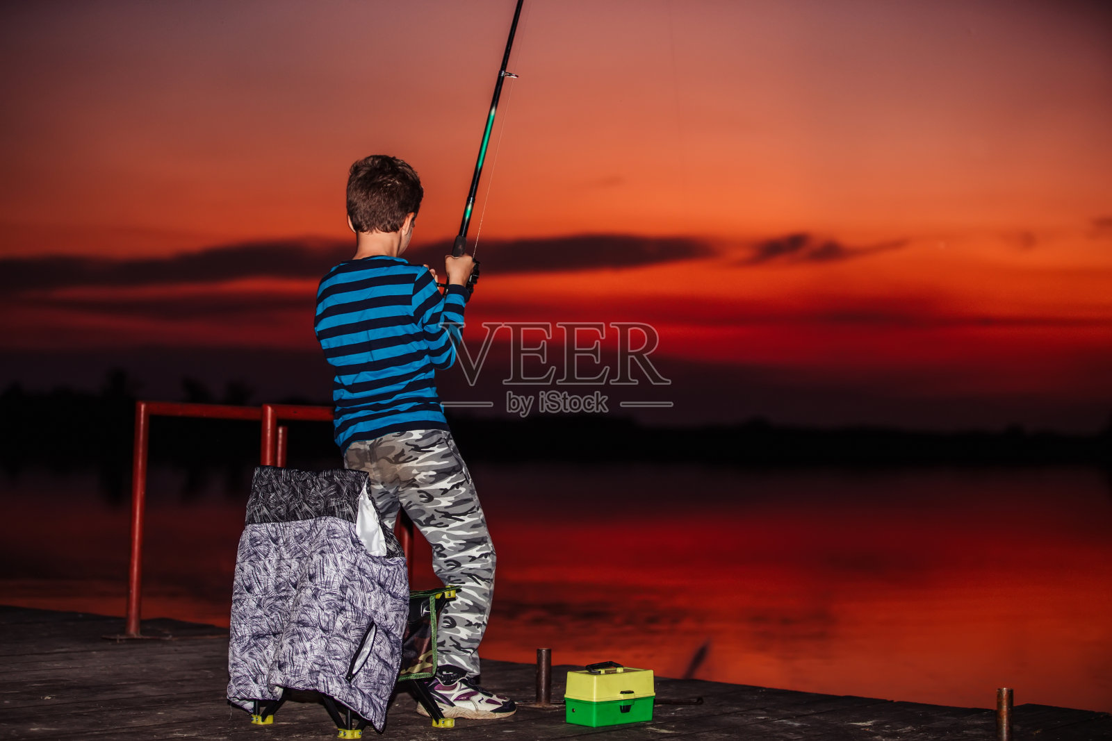 日落时分，可爱的小男孩在湖边钓鱼。照片摄影图片