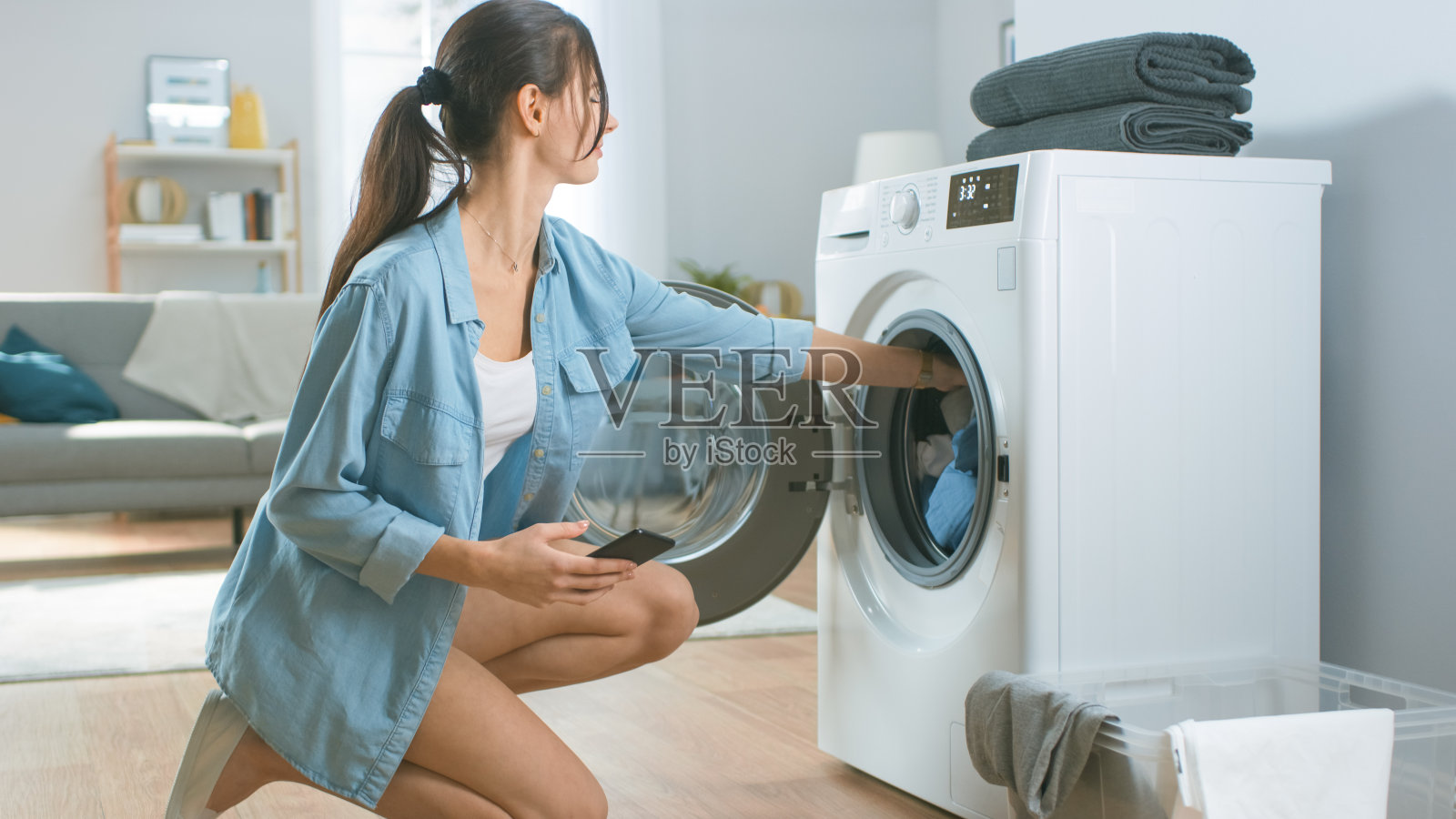 美丽的年轻女子坐在洗衣机旁边的膝盖上。她在洗衣机里装了脏衣服，并用智能手机配置了洗衣机。拍摄于客厅与现代室内。照片摄影图片