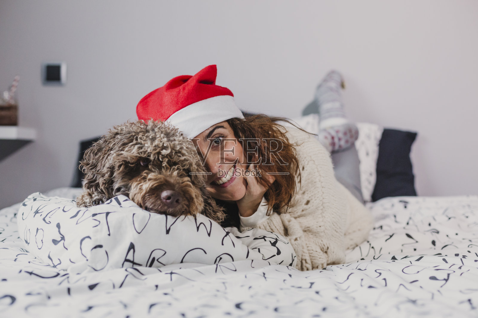 年轻女子在家里戴着圣诞老人的帽子和她的狗在床上玩。在室内。圣诞节的概念照片摄影图片
