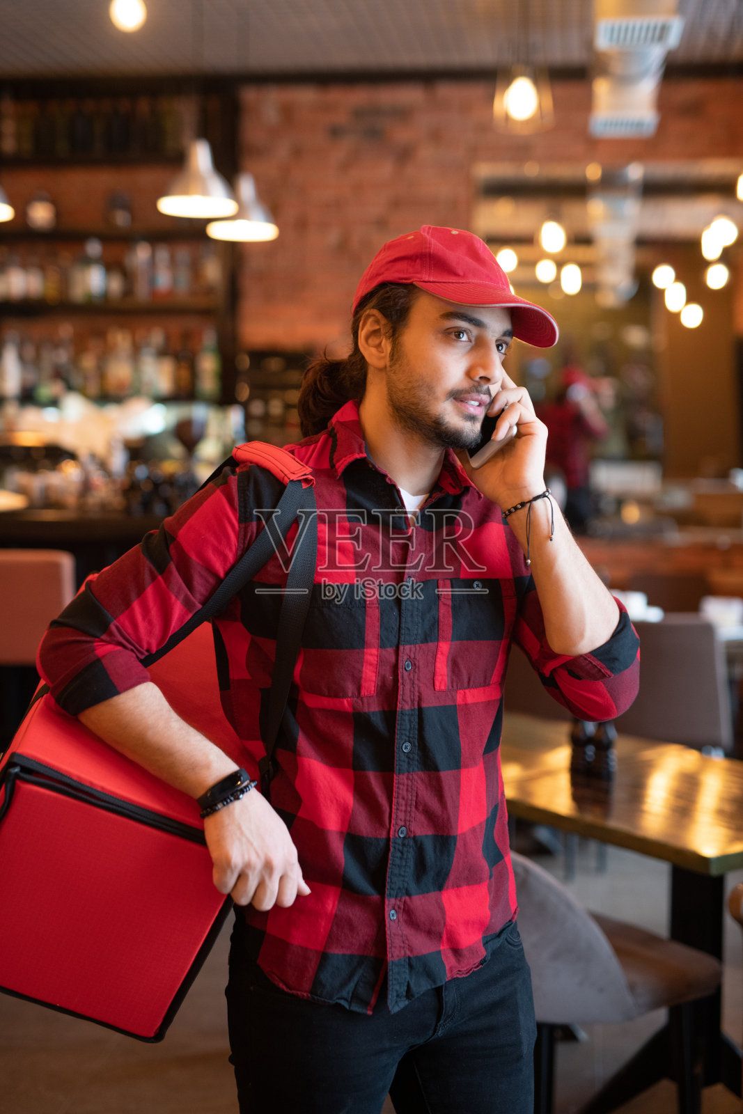 肩上扛着一个大红色袋子的年轻快递员正在咖啡馆给客户打电话照片摄影图片