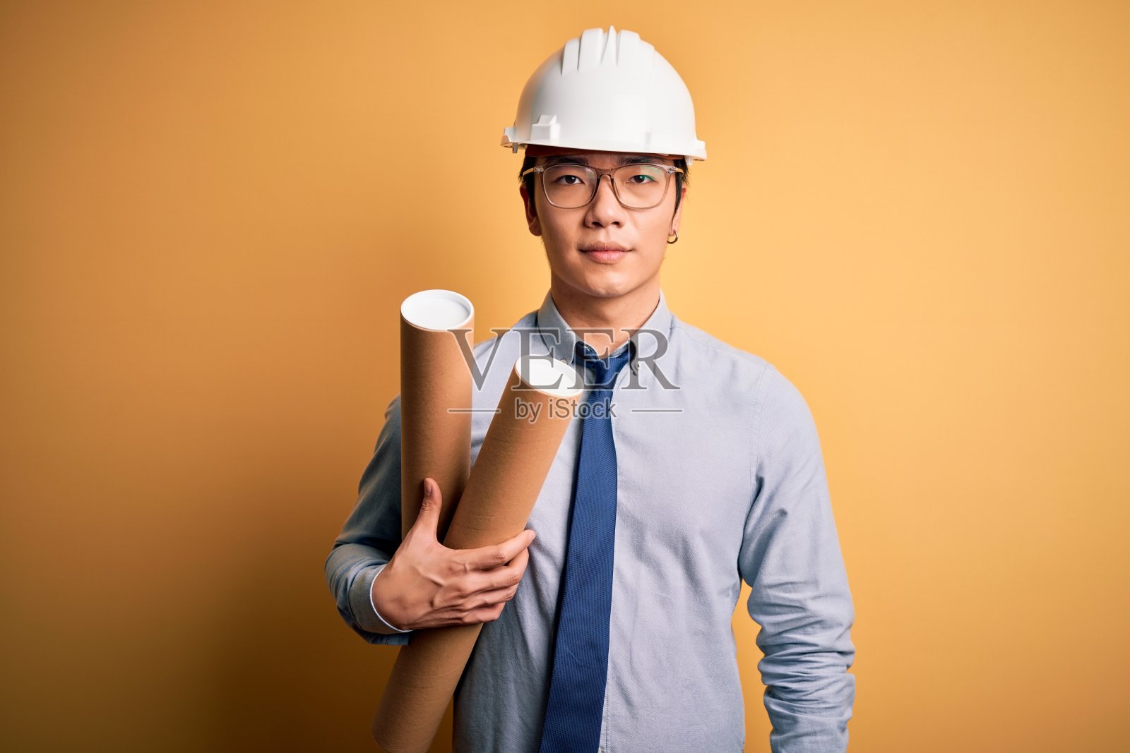 年轻英俊的中国建筑师戴着眼镜和安全帽拿着蓝图，聪明的脸上有自信的表情，思考认真照片摄影图片