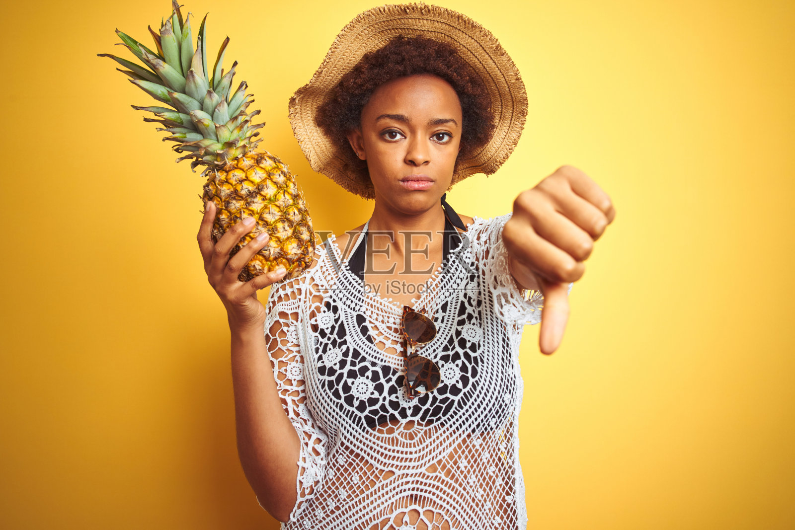年轻的非洲裔美国妇女与非洲式的头发，举着菠萝在黄色孤立的背景，愤怒的脸，消极的迹象显示不喜欢拇指下来，拒绝的概念照片摄影图片