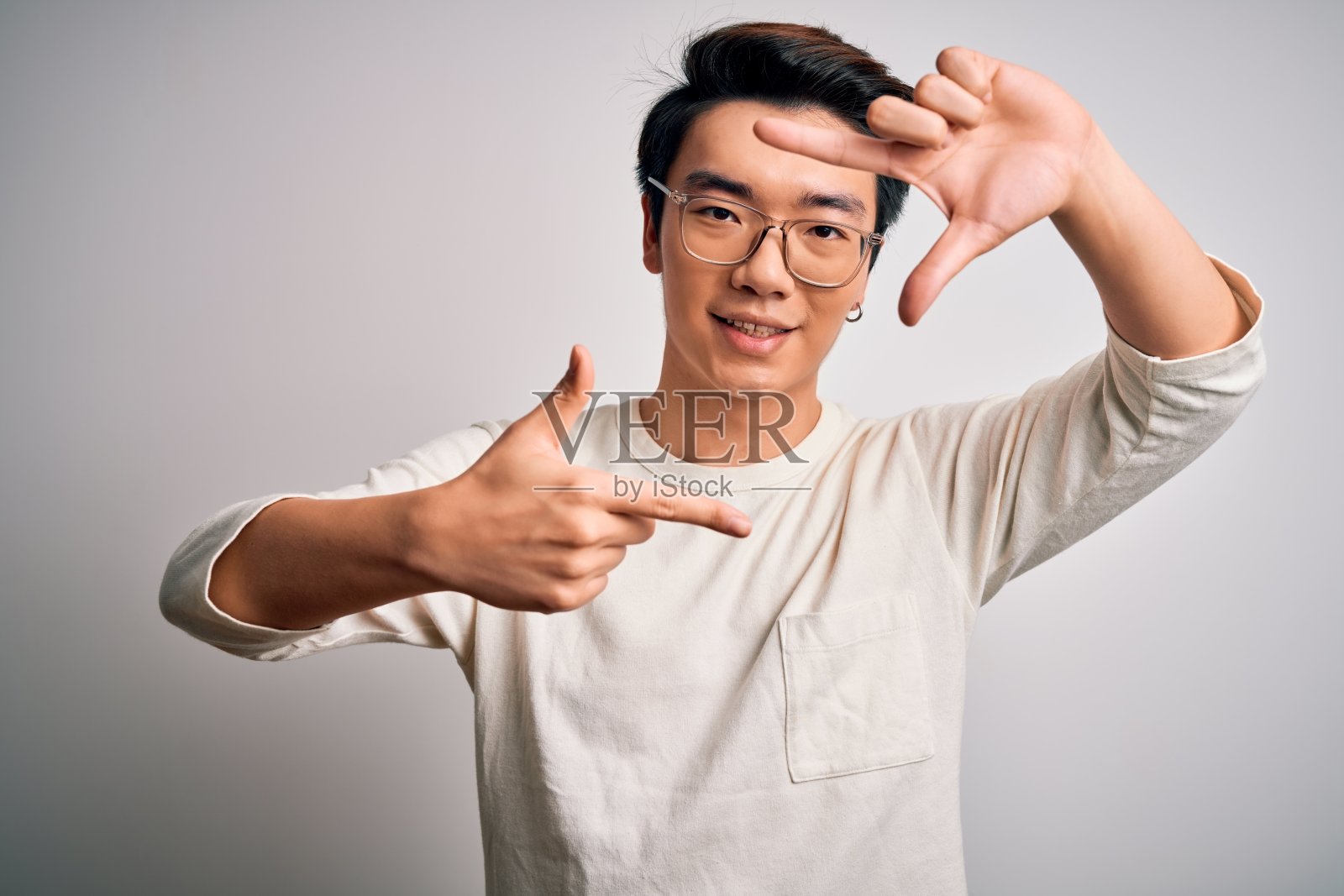 年轻英俊的中国男子穿着休闲t恤和眼镜在白色的背景，微笑的框架与手和手指与快乐的脸。创意和摄影概念。照片摄影图片