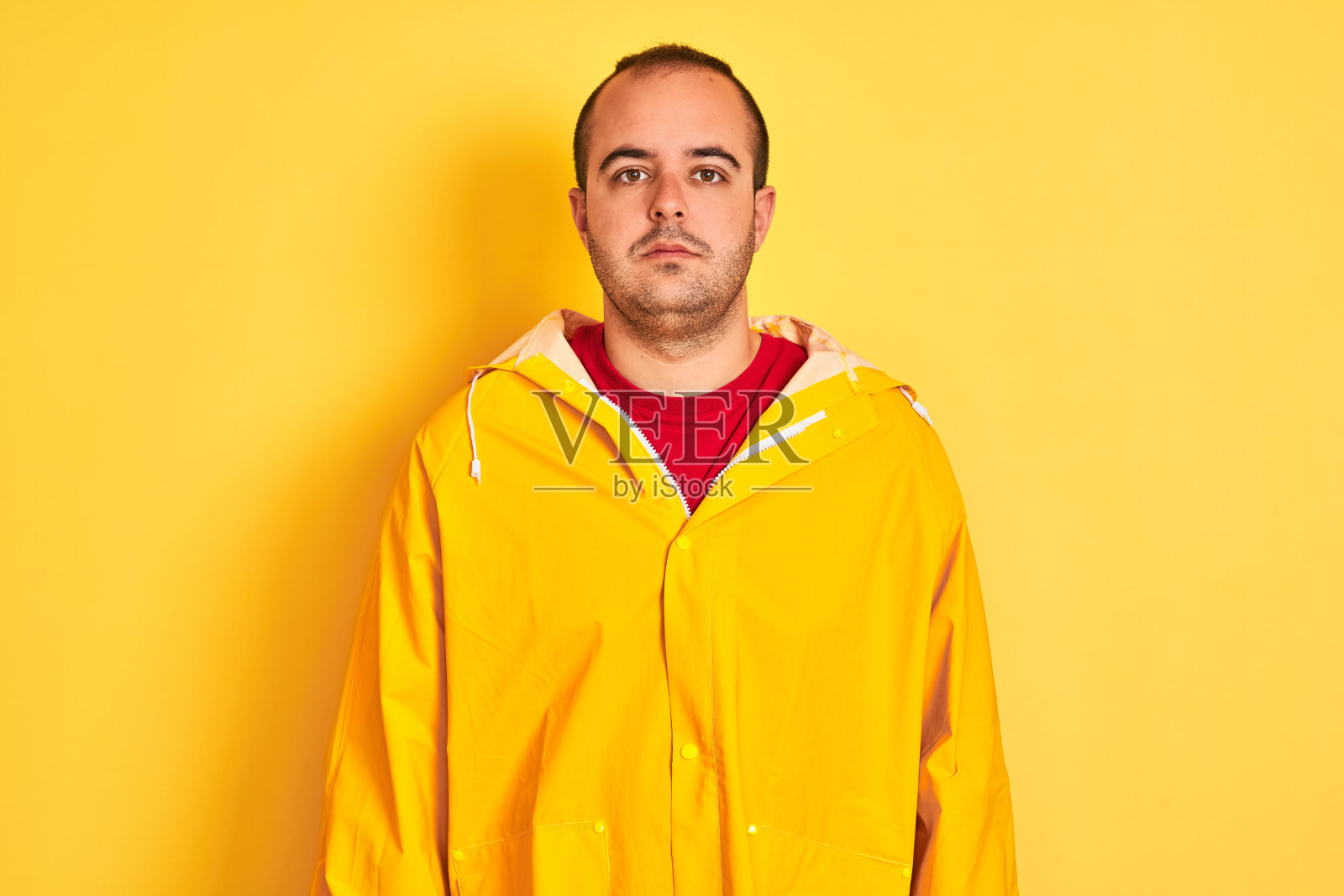 一个年轻人穿着雨衣站在孤立的黄色背景上，神情轻松，一脸严肃。简单自然的看相机。照片摄影图片