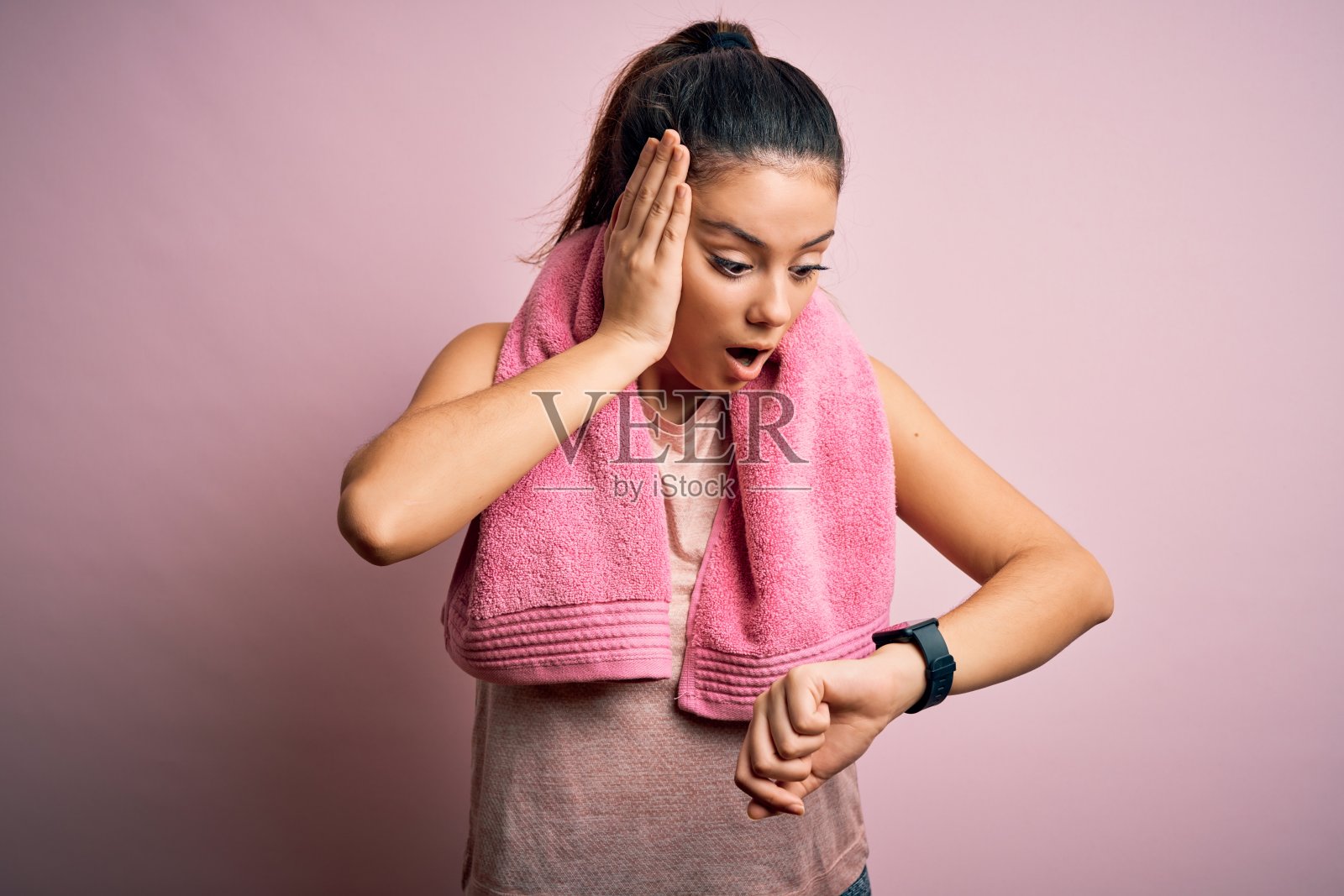 年轻漂亮的黑发女运动员穿着运动服和毛巾在粉红色的背景下看着手表时间着急，害怕迟到照片摄影图片