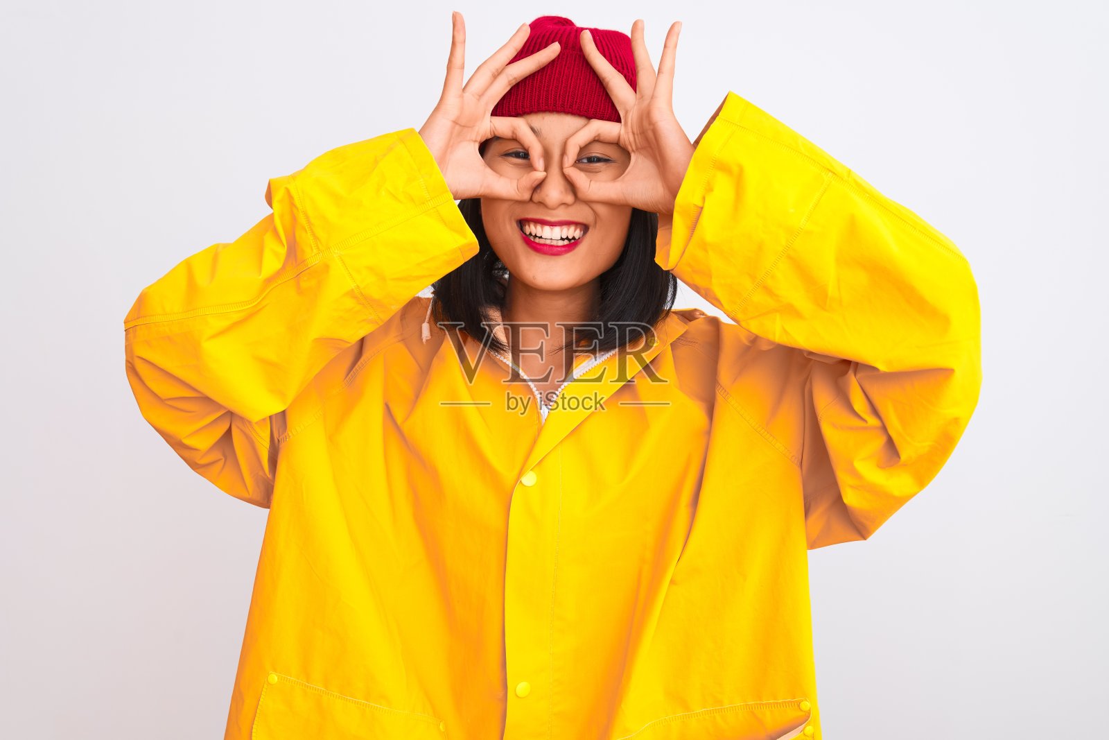 年轻美丽的中国女人穿着雨衣和羊毛帽，在孤立的白色背景下做着不错的手势，就像望远镜伸出舌头，眼睛透过手指看。疯狂的表情。照片摄影图片