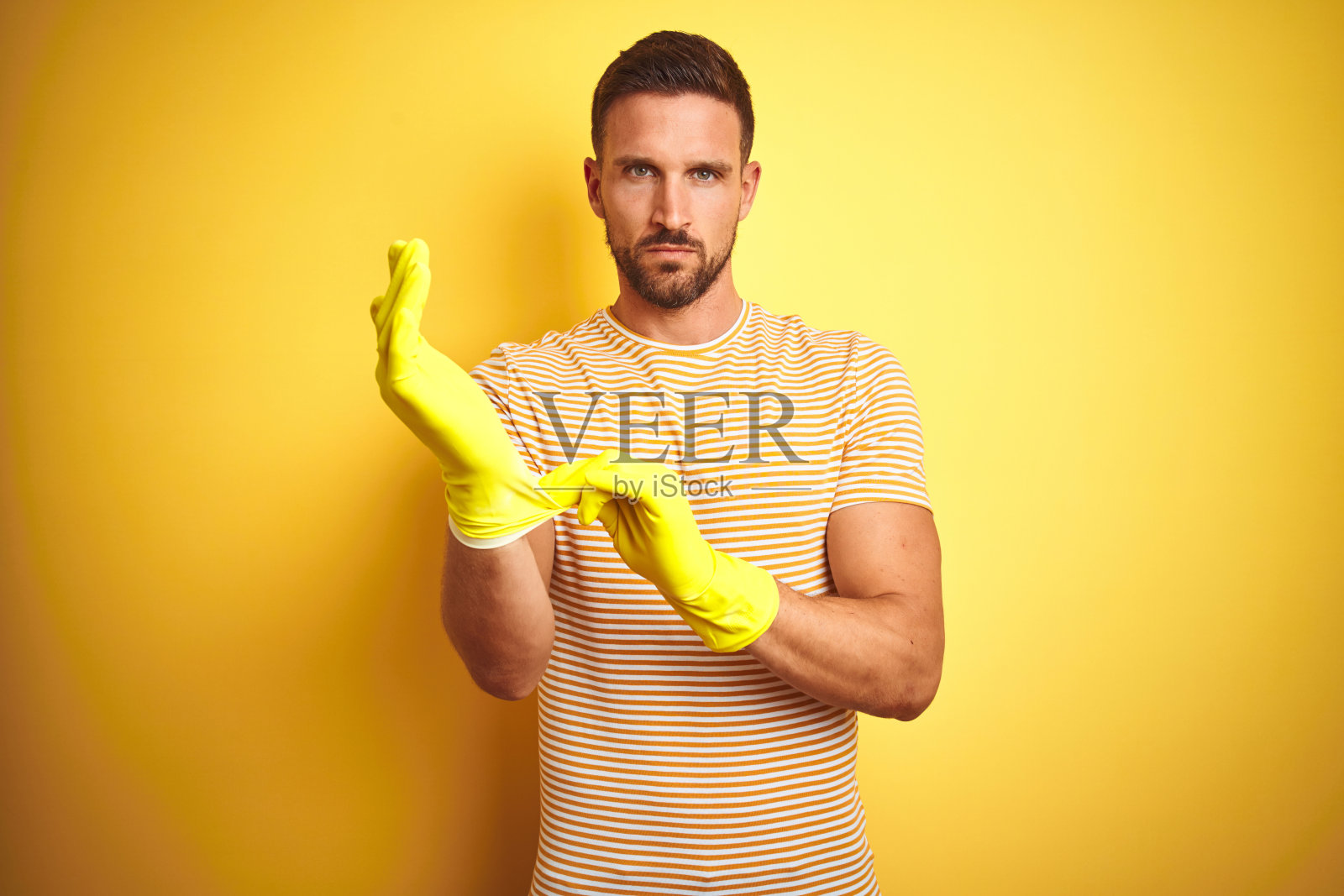 年轻英俊的男子戴着清洁手套，为家务孤立黄色的背景与自信的表情，聪明的脸上思考认真照片摄影图片