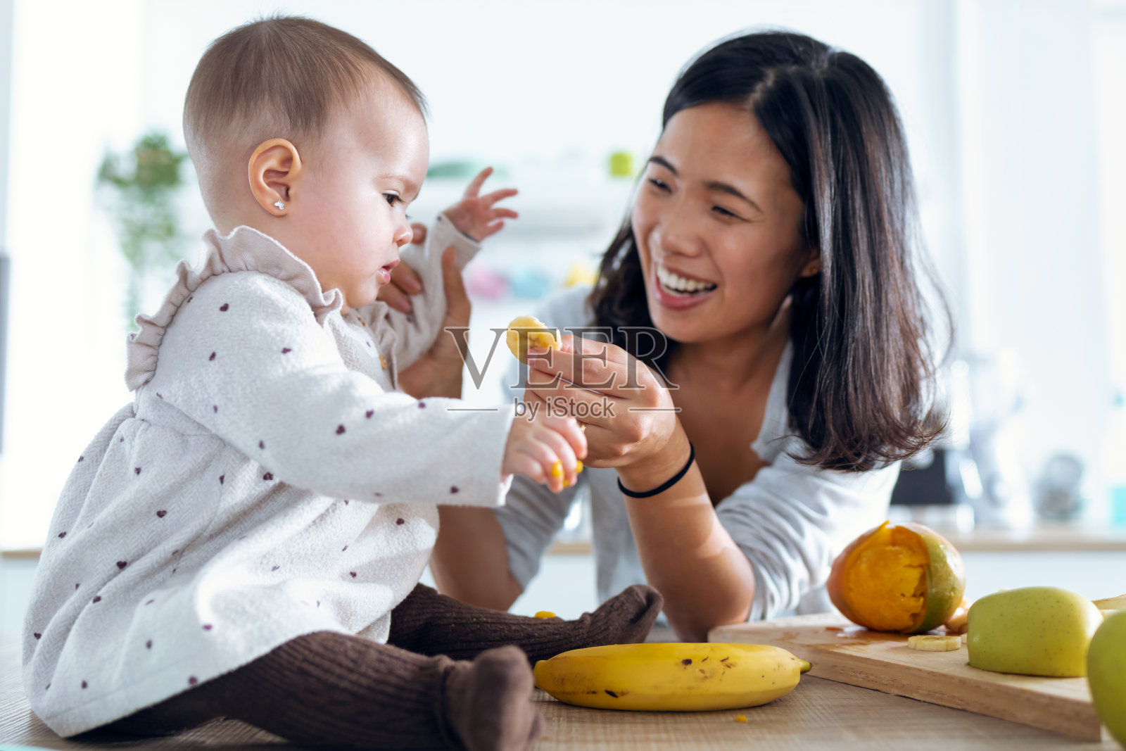 快乐的年轻妈妈在家里的厨房里用香蕉喂她可爱的小女孩。照片摄影图片