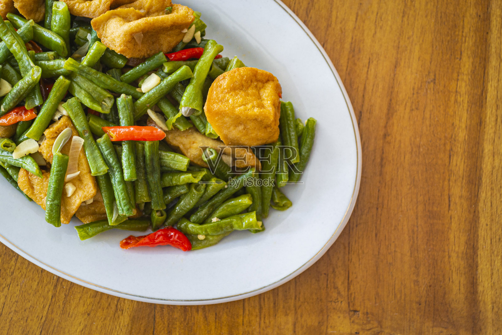 Tumis tahu kacang panjang是印度尼西亚的一种食物，由新鲜的长豆、豆腐、辣椒和其他原料煸炒而成。在木制背景上的白色盘子里照片摄影图片
