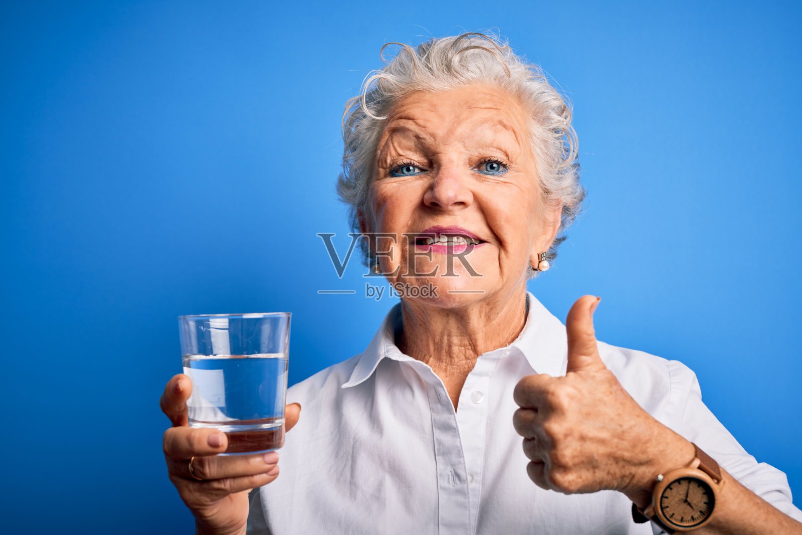 资深美丽的女人喝杯水站在孤立的蓝色背景开心地笑着做ok的手势，用手指竖起大拇指，优秀的手势照片摄影图片