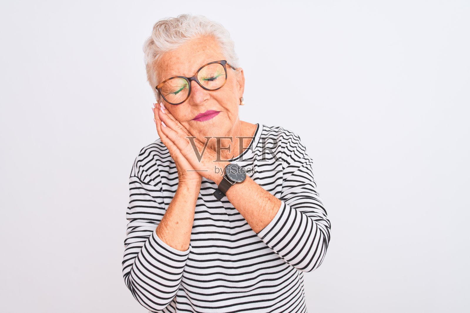 一位白发苍苍的老妇人戴着条纹海军蓝t恤，背景是孤立的白色，她疲倦地睡觉，做着梦，闭着眼睛微笑着双手合十。照片摄影图片