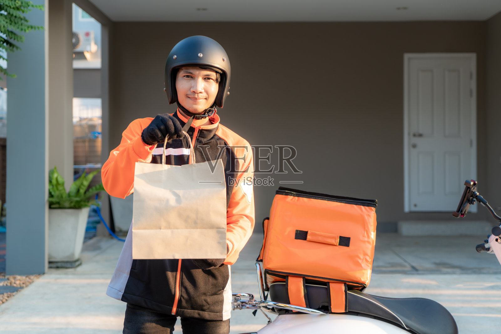 外卖亚洲男子身穿橙色制服，准备送外卖食品袋在顾客家门口，食品盒放在踏板车上，快递食品和网上购物概念。照片摄影图片