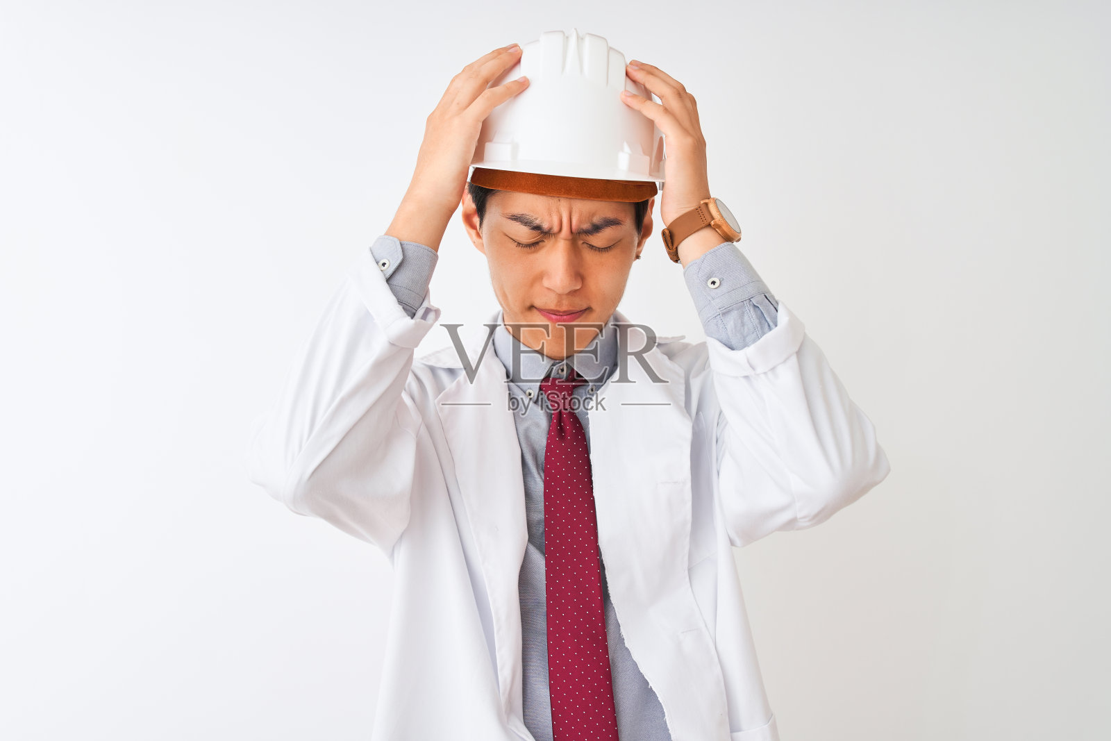 一名中国建筑师穿着大衣，戴着头盔，站在孤立的白色背景下，饱受头痛之苦，因为疼痛和偏头痛而绝望和紧张。手放在头上。照片摄影图片